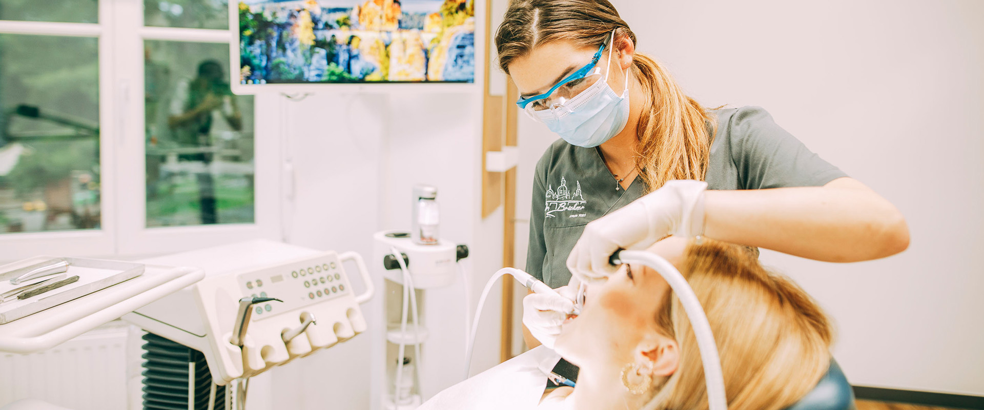 Professionelle Zahnreinigung im IMplantatzentrum Brückner: auf dem Bild sehen wir eine Patientin im Stuhl und eine Prophylaxe-ZFA. Die jungen blonde ZFA reinigt die Zähne der Patientin.