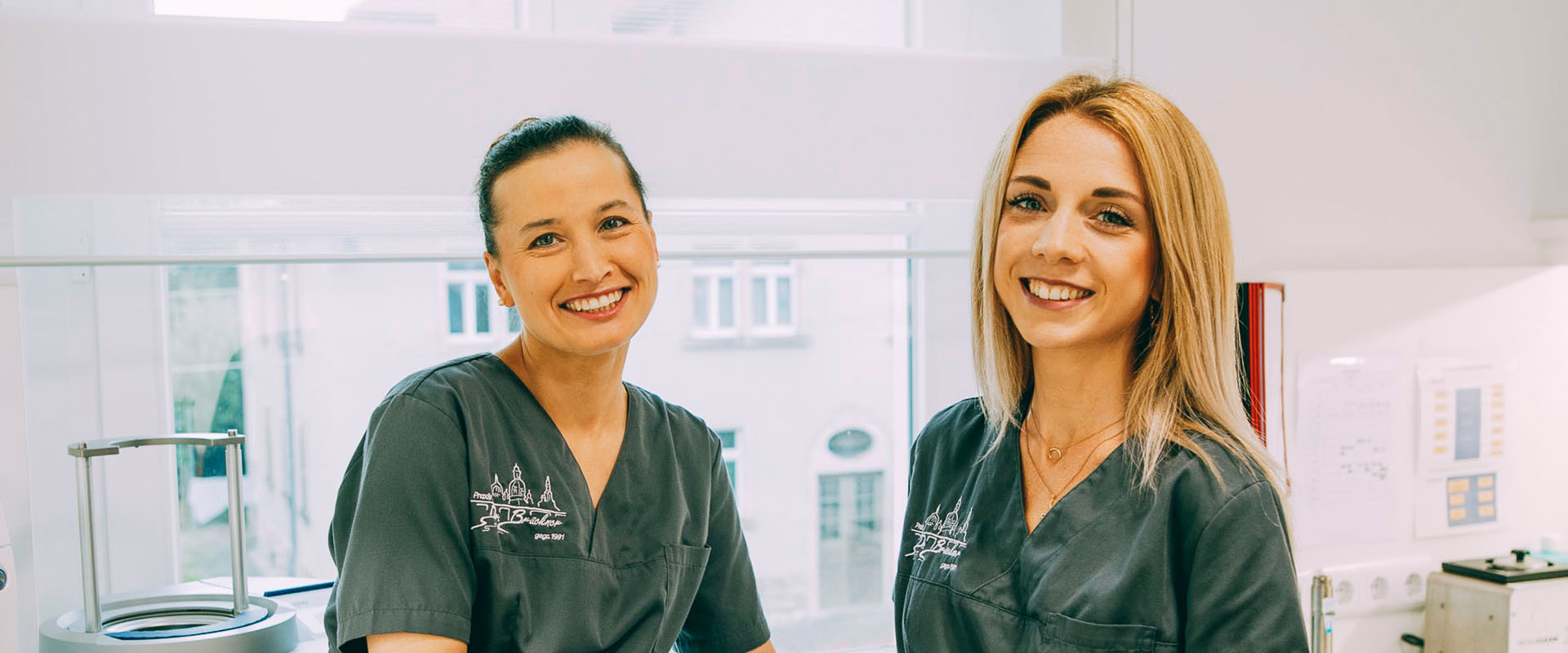 Zwei blonde Frauen lächenln in die Kamera. Die jungen, attraktiven Damen sind das Bleaching-Team der Zahnarztpraxis Brückner. Sie hellen gekonnt und professionell die Zähne auf.