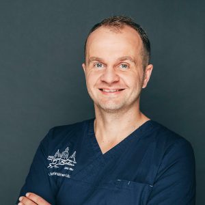 Zahnarzt in blauer Arbeitskleidung lächelt freundlich in die Kamera